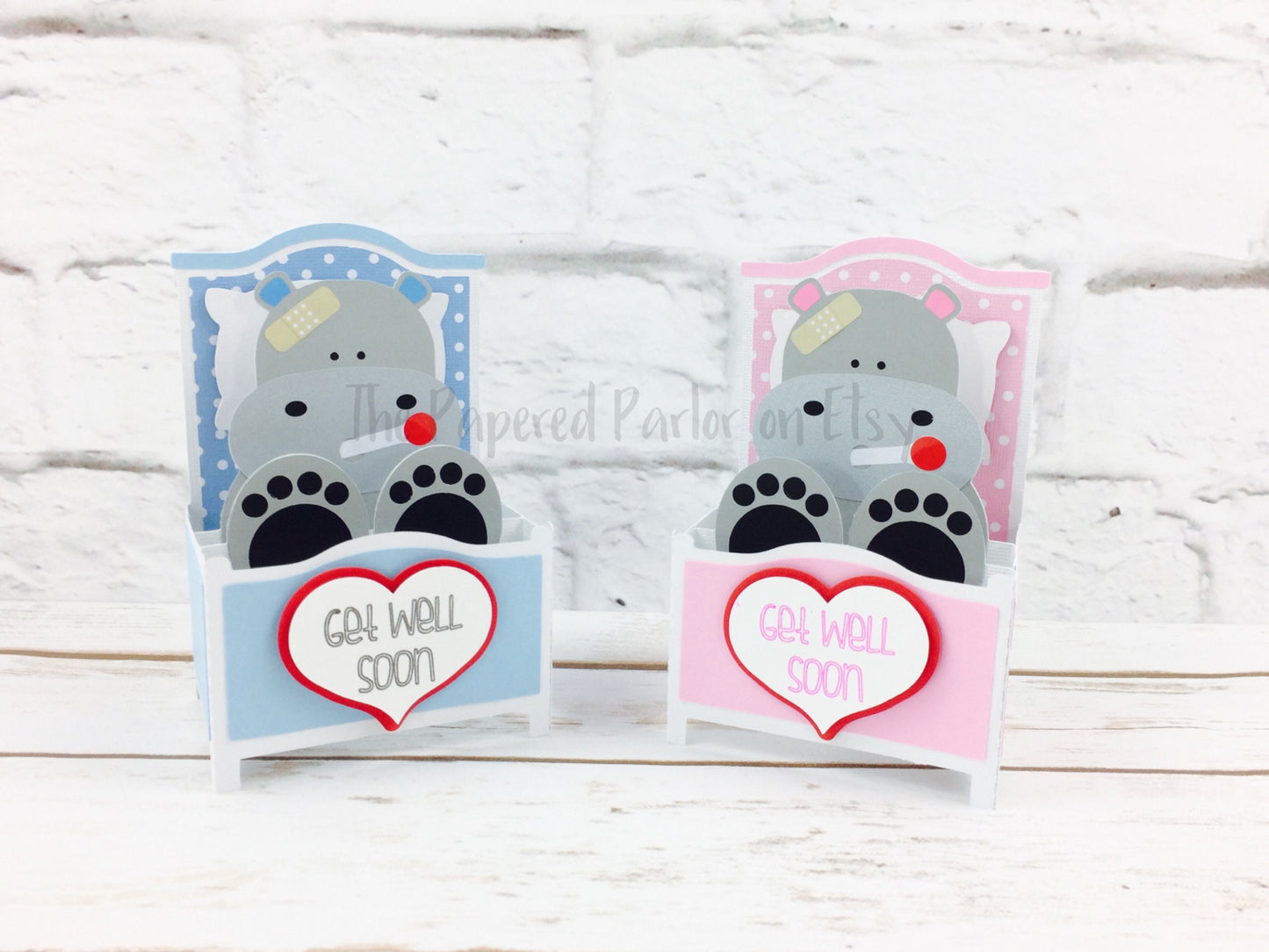 hippo get well soon pop up box card. a boy hippo is in a light blue bed while the girl hippo is in a pink bed. both have a bandage on their foreheads and a thermometer in their mouths. on the end of the bed is a heart that says get well soon.